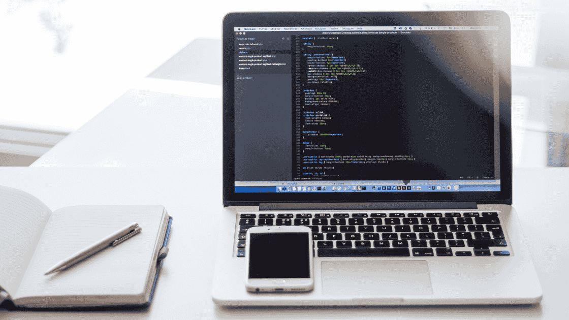 Office desk with notepad, pencil, laptop with code on the screen and a phone on the laptop