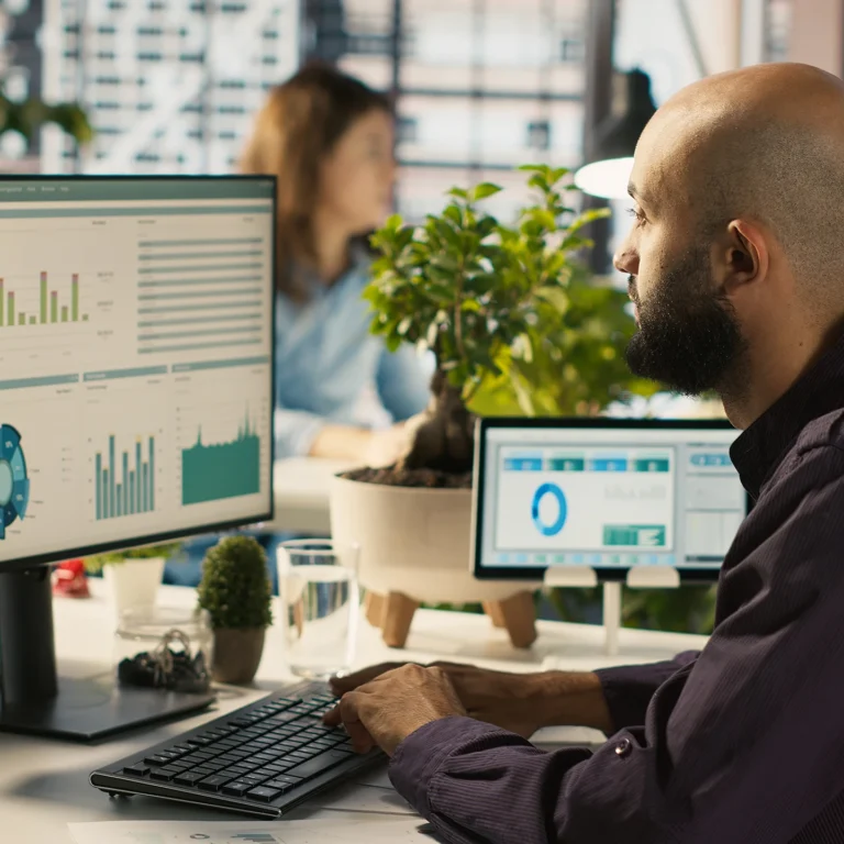 A man at a desk with a computer, while a woman sits behind him, both engaged in data on their screens.