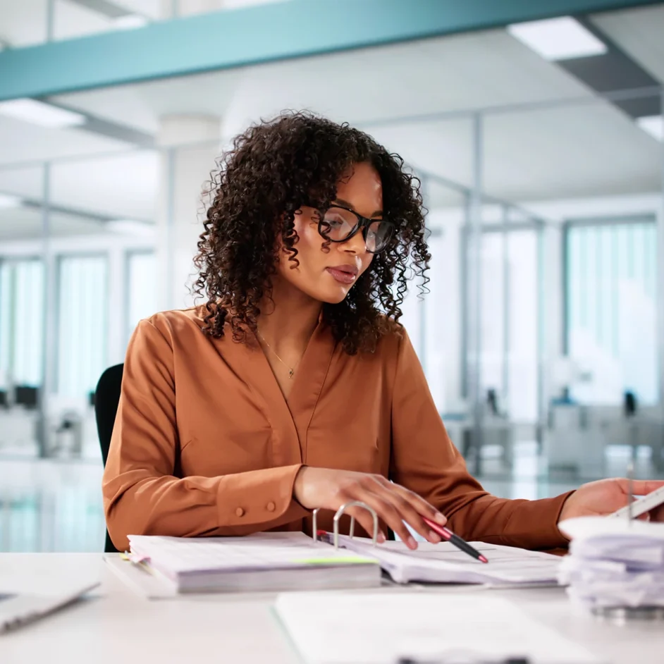 A woman wearing glasses sits at a desk, focused on her laptop, surrounded by books and a notepad.