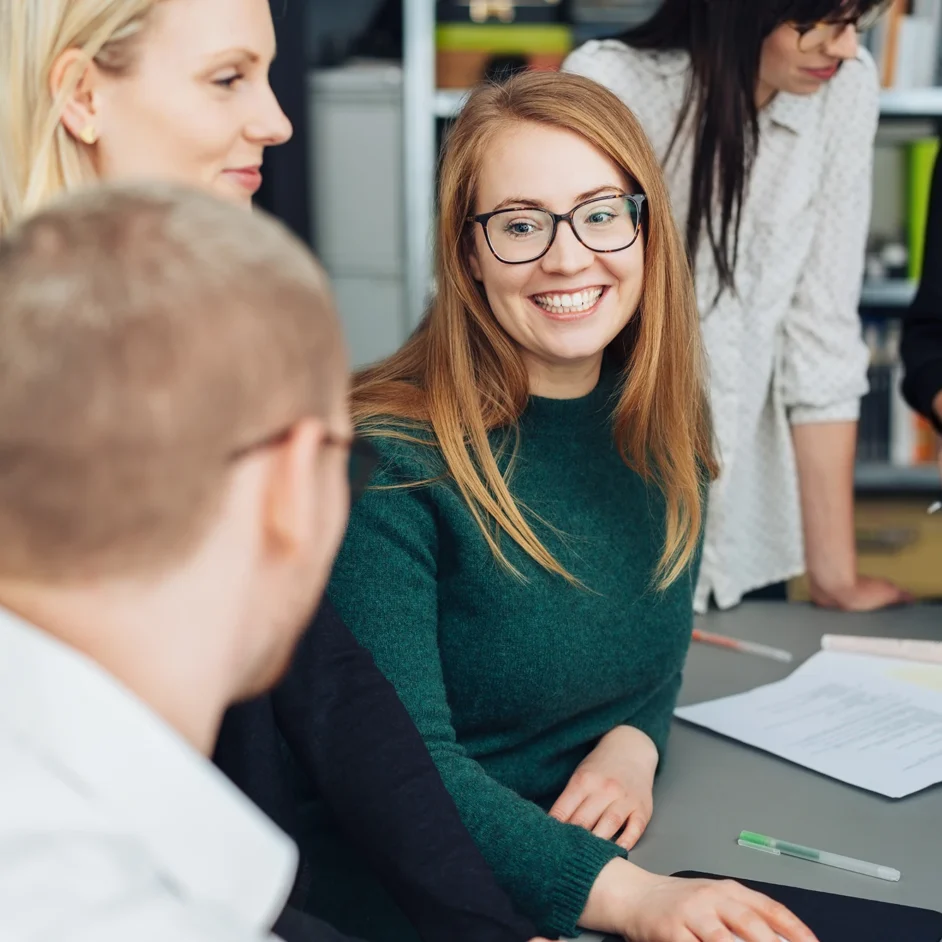 A group of people sitting around a table, with a smiling woman at the centre, enjoying a friendly conversation.