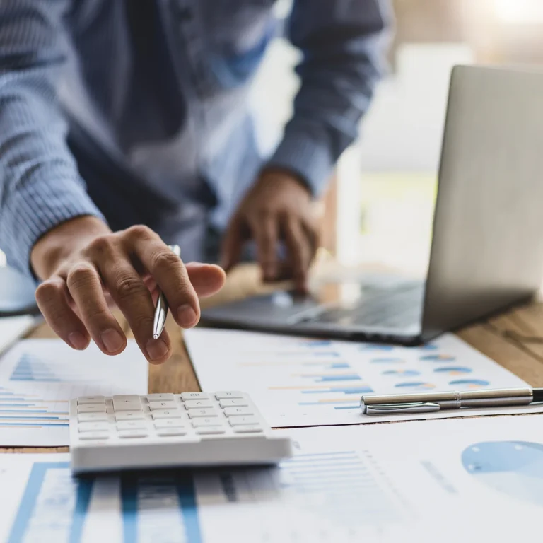 A person calculates on a desk with papers and a laptop, focusing intently on their work.