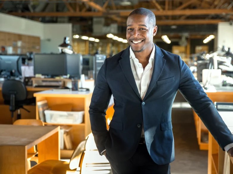 A man in a suit stands confidently in a modern office, surrounded by sleek furniture and large windows.