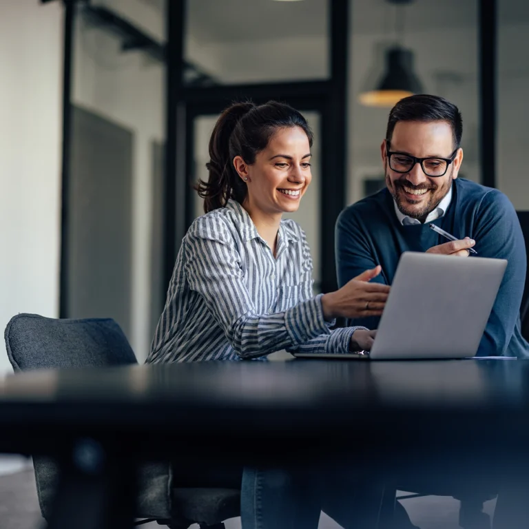 A man and woman sit at a table, focused on a laptop, engaged in conversation or collaboration.