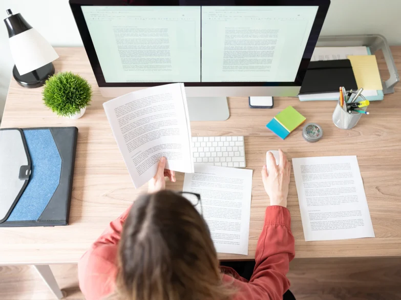 A woman sits at a desk with a computer and papers, focused on her work in a bright, organised workspace.
