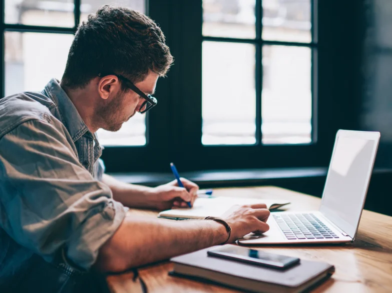 A man wearing glasses is focused on working on his laptop at a desk.