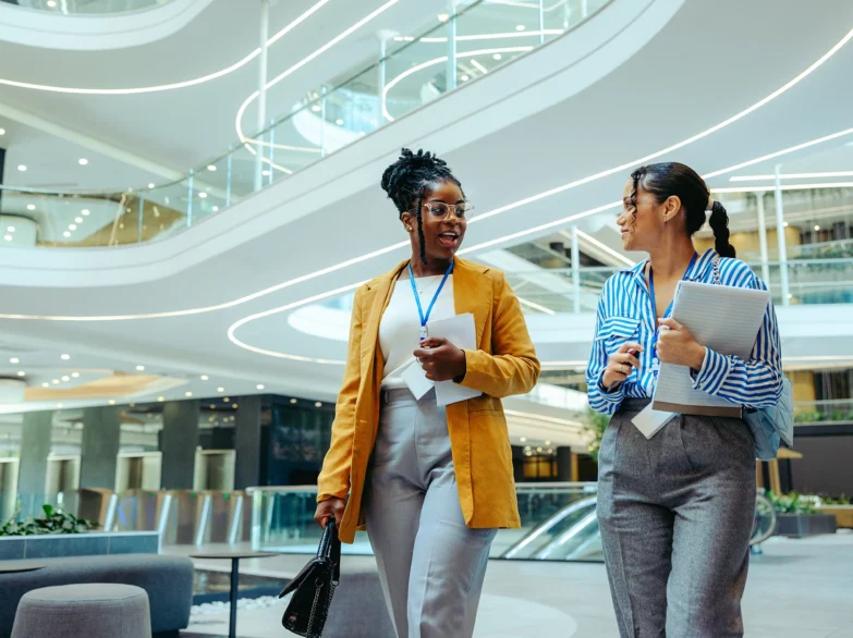 Two women walking together in a modern office building, engaged in conversation.