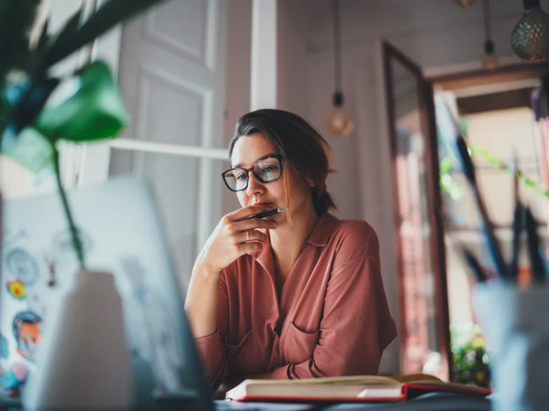 A woman wearing glasses sits at a desk, focused on her laptop while working.