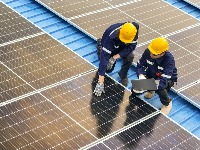 Two men in hard hats installing solar panels on a rooftop under a clear blue sky.