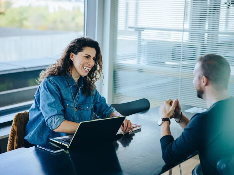 A man and woman sit at a table, focused on a laptop, engaged in conversation.