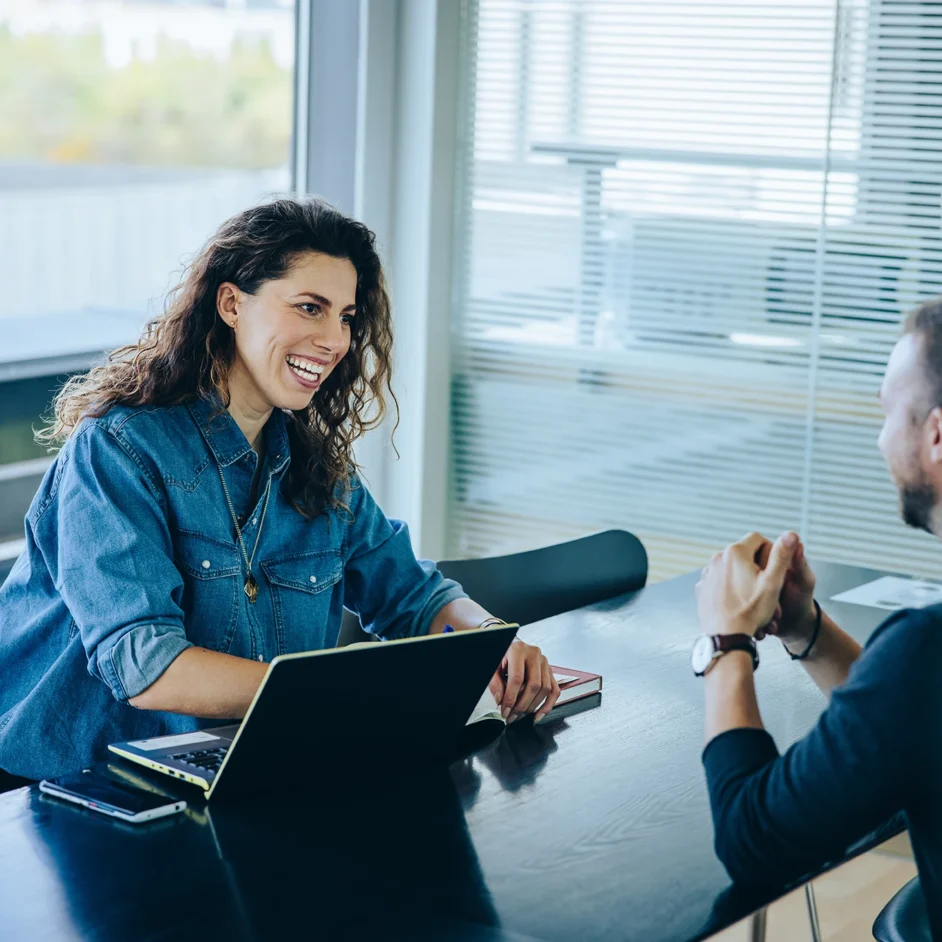 A man and woman sit at a table, focused on a laptop, engaged in conversation.