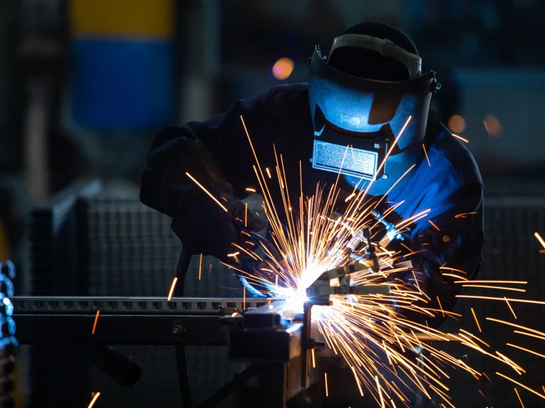 A man welding in a factory, surrounded by flying sparks and industrial equipment.