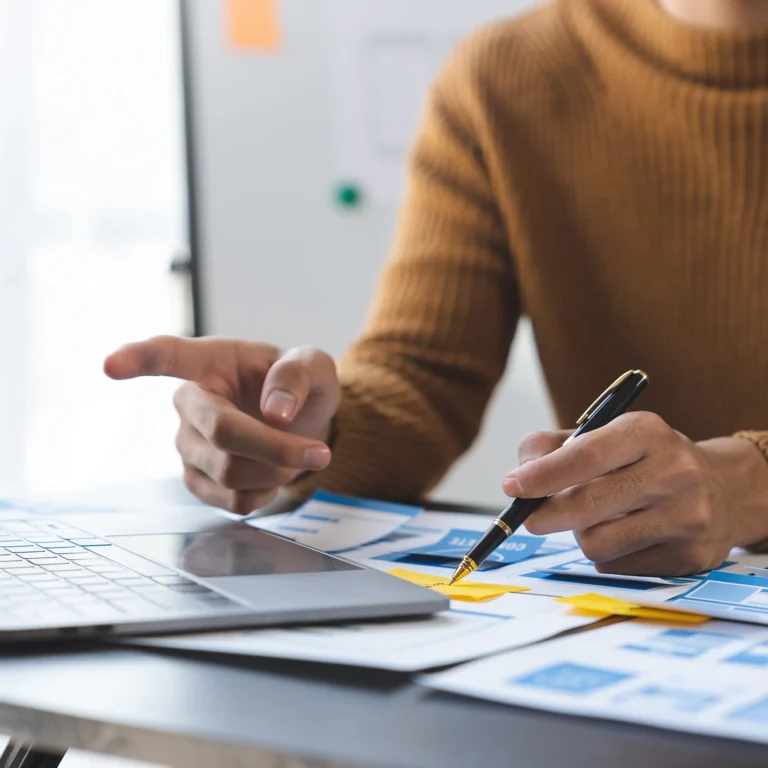 Two people collaborating on a laptop, surrounded by papers and pens, focused on their work.