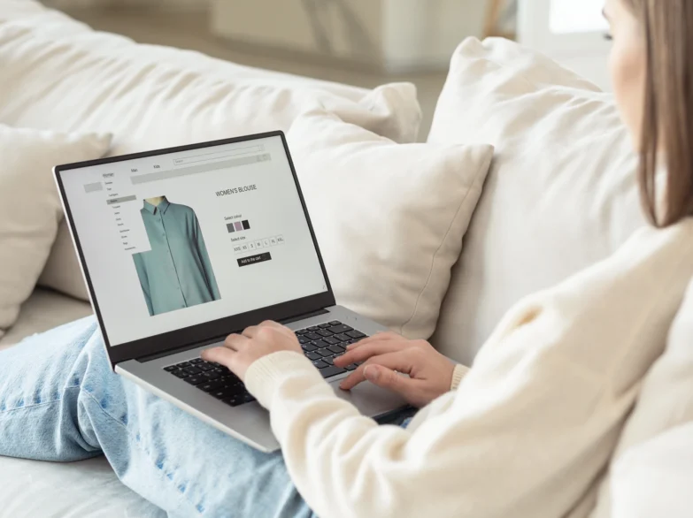 A woman sits on a couch, wearing a shirt and shopping on a laptop.