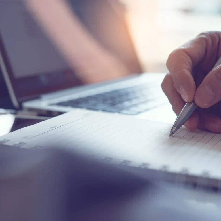 A man writes in a notebook with a pen, sitting at a desk filled with papers and a cup of coffee.