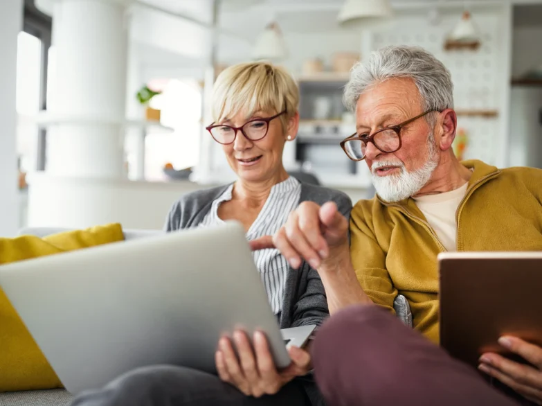 An older couple sitting together, smiling while using a laptop at home.