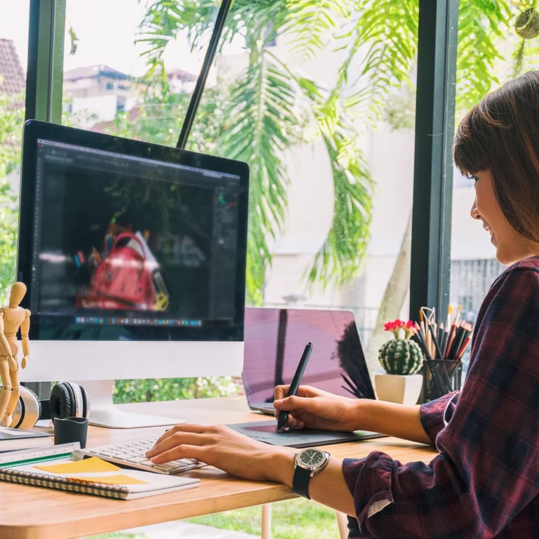 A woman works on her computer at a desk, with natural light streaming in through a nearby window.