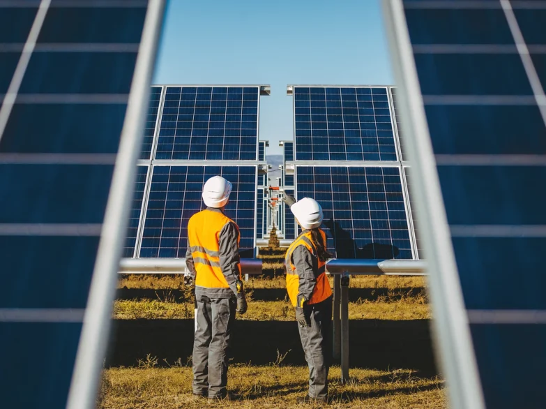 Two people in orange vests stand in front of solar panels, showcasing renewable energy efforts in a sunny environment.