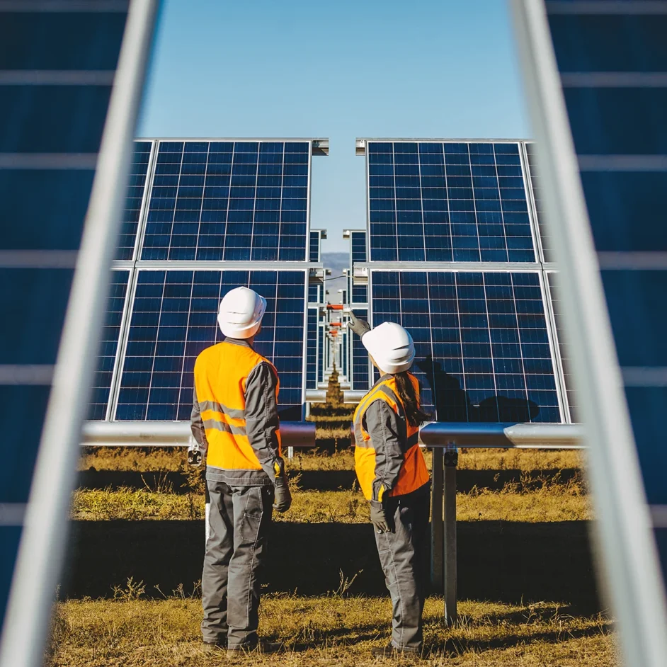 Two people in orange vests stand in front of solar panels, showcasing renewable energy efforts in a sunny environment.