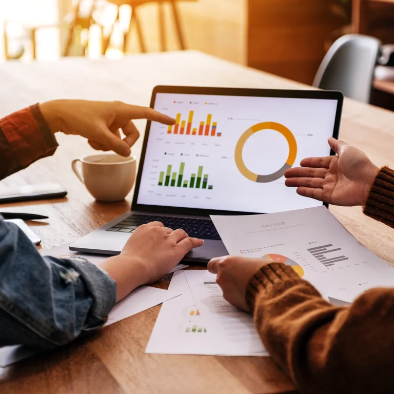 Two people seated at a table, working together with a laptop and scattered papers.