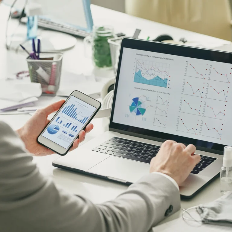 A man analyses graphs on his laptop while using his phone for additional data.